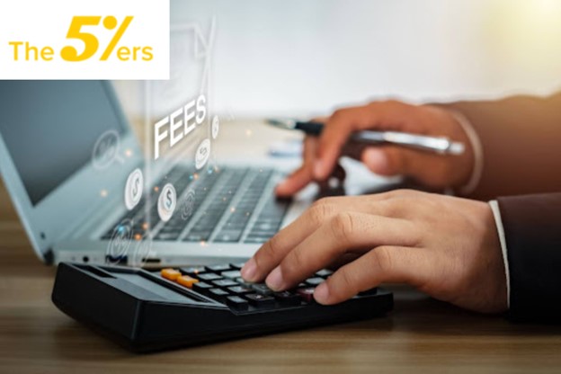 Hands using a calculator and laptop with 'Fees' displayed, highlighting high fees and hidden costs at The5ers trading firm.”